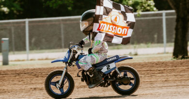 Little kid riding his motorcycle on the dirt track while holding a checkered flag with the Mission Foods logo on it.