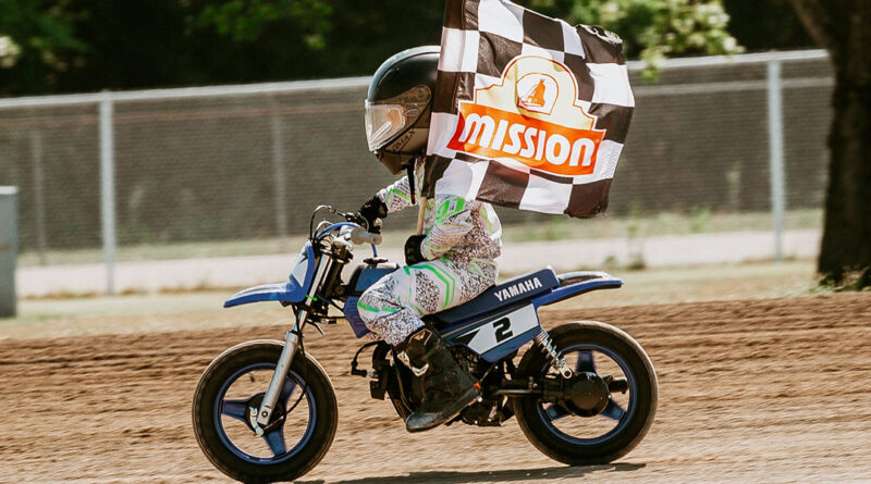 Little kid riding his motorcycle on the dirt track while holding a checkered flag with the Mission Foods logo on it.