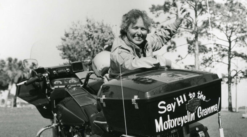 A black and white photo of Hazel Kolb leaning against her motorcycle which has "say hi to the Motorcycling' Granma" on that back of it